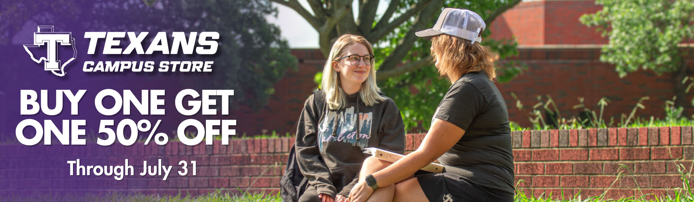 Two people sit on grass in front of a brick wall with a 